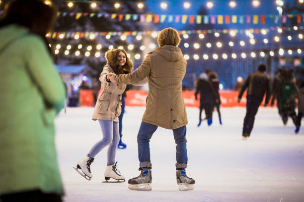 young couple in love caucasian man with blond hair with long hair and beard and beautiful woman have fun, active date skating on ice scene in town square in winter on christmas eve - outdoor skating rink stock pictures, royalty-free photos & images