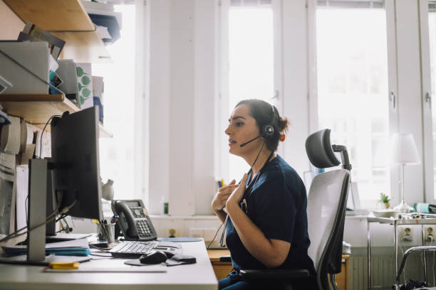 side view of mature female nurse doing video call while sitting on chair in clinic - telehealth stock pictures, royalty-free photos & images