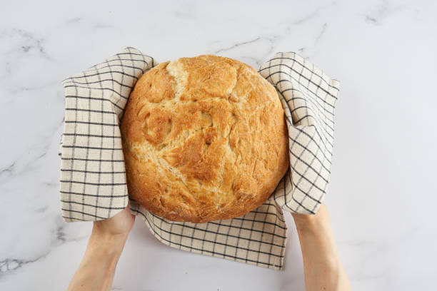 hands holding whole loaf of freshly baked white wheat bread on linen napkin on home kitchen table - round bread loaf stock pictures, royalty-free photos & images